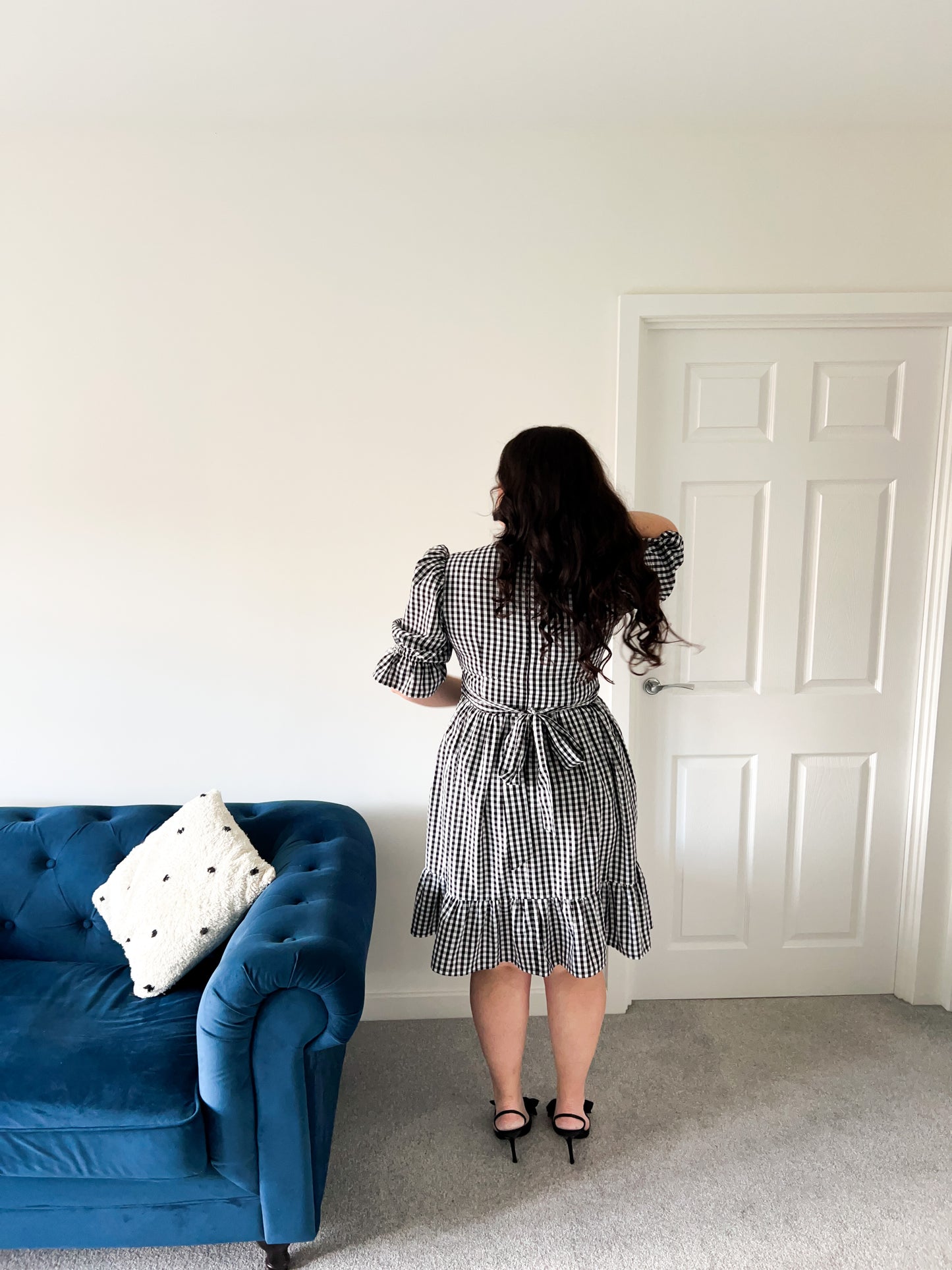 Woman in a checkered dress standing in a room with a blue sofa and white door.